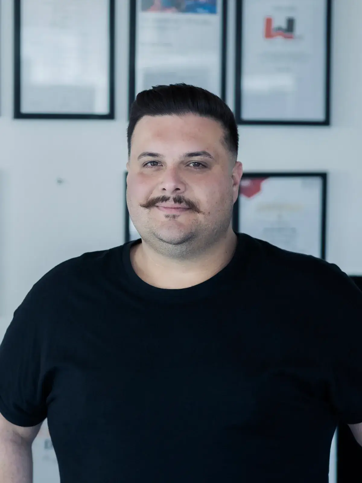 Okan Aydin, Kfz-Sachverständiger bei 5Expert, vor seiner Wand mit Zertifikaten und Fortbildungsnachweisen